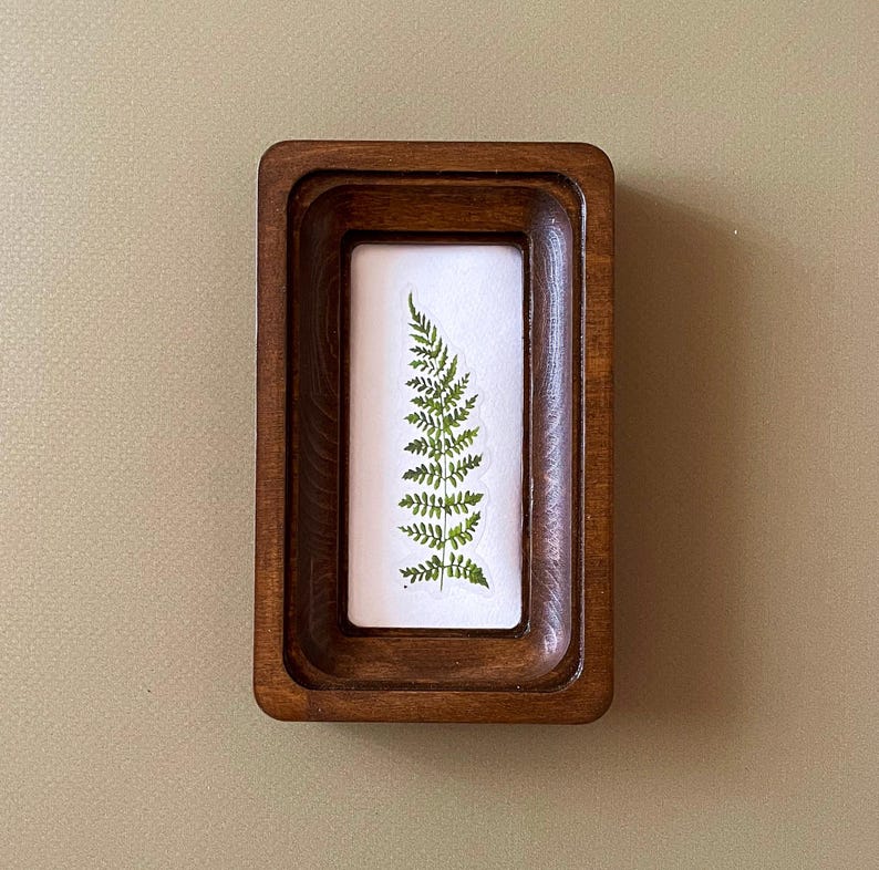 May include: A rectangular wooden tray with a recessed interior holds a white rectangle with a pressed fern. The fern is green and detailed, showcasing the plant's delicate fronds. The tray has a dark brown finish.