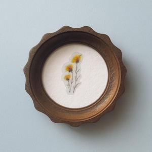 May include: A round, wooden framed art piece featuring a botanical illustration of yellow wildflowers. The frame has a scalloped edge and a warm brown finish. The artwork is centered on a white background.