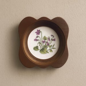 May include: A circular framed artwork with a wooden frame. The image is a botanical illustration of purple violets with green leaves on a white background. The frame has a scalloped edge and is made of dark brown wood.