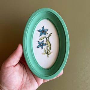 May include: An oval, mint green picture frame displaying a floral illustration. The artwork shows two blue flowers with green leaves and stems on a white background. The frame is held by a hand.