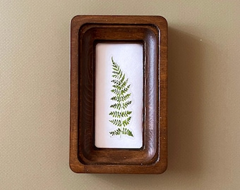 May include: A rectangular wooden tray with a recessed interior holds a white rectangle with a pressed fern. The fern is green and detailed, showcasing the plant's delicate fronds. The tray has a dark brown finish.