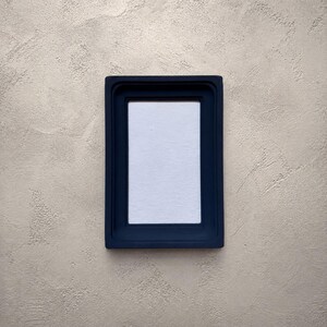 Pode incluir: Uma moldura retangular com acabamento azul-marinho mate e um design de borda lisa e escalonada. A moldura apresenta uma orientação vertical limpa e envolve uma inserção central retangular branca e lisa para exibir fotografias.
