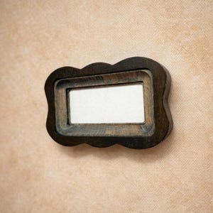 May include: A dark brown wooden frame with a scalloped edge and a rectangular cutout. The frame features a visible wood grain texture and surrounds a plain, off-white rectangular center panel, creating a layered, dimensional appearance.