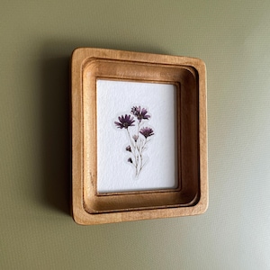 以下が含まれることがあります： 小さな長方形の木製フレームに、押し花のアート作品が飾られています。 フレームは明るい茶色です。 アートワークは、白い背景に濃い紫色の花のクラスターを特徴としています。