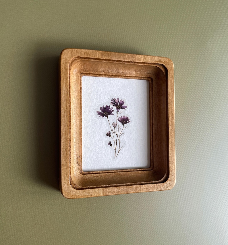 以下が含まれることがあります： 小さな長方形の木製フレームに、押し花のアート作品が飾られています。 フレームは明るい茶色です。 アートワークは、白い背景に濃い紫色の花のクラスターを特徴としています。