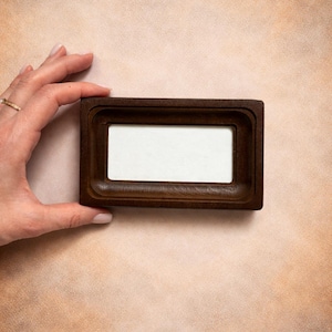 May include: A rectangular dark brown wooden tray featuring a smooth, recessed center with a white rectangular insert. The tray has a solid, thick frame with rounded inner edges and a flat outer border, presenting a clean and functional design.