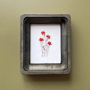 Pode incluir: Uma obra de arte emoldurada com um grupo de flores vermelhas sobre um fundo branco. A obra está envolta em uma moldura de madeira retangular cinza escuro. O fundo é verde-oliva suave.