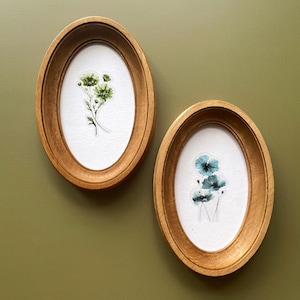 Puede incluir: Dos marcos ovalados de madera con arte botánico. Uno presenta flores verdes, el otro flores azules. Los marcos están montados en una pared verde, creando una exhibición natural y artística.