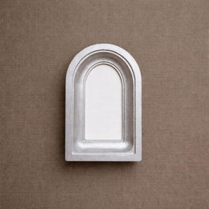 Könnte beinhalten: Ein silberfarbener, bogenförmiger Rahmen mit einer vertieften, glatten, cremeweißen Mittelplatte. Der Rahmen verfügt über ein gestuftes, geschichtetes Randdesign, das Tiefe erzeugt, mit einer flachen Basis und einer abgerundeten Oberkante, was eine klare, solide Struktur ergibt.