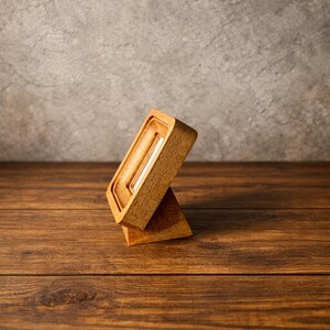 May include: A wooden soap dish with a rectangular design, featuring a raised edge and a slatted interior for drainage. The dish is supported by a triangular base, showcasing the natural wood grain. The background is a textured gray wall and a wooden surface.