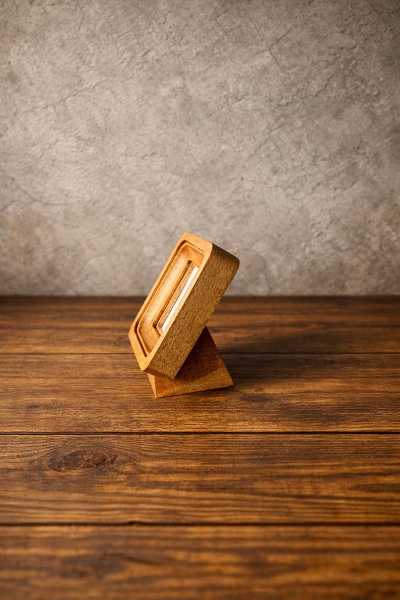 May include: A wooden soap dish with a rectangular design, featuring a raised edge and a slatted interior for drainage. The dish is supported by a triangular base, showcasing the natural wood grain. The background is a textured gray wall and a wooden surface.