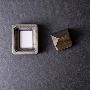 Puede incluir: Un peque&ntilde;o marco de fotos cuadrado de madera con un inserto blanco y un bloque de madera geom&eacute;trico. El marco tiene un acabado gris y un &aacute;rea empotrada para la foto. El bloque es de color marr&oacute;n oscuro y tiene un dise&ntilde;o facetado. Los objetos est&aacute;n sobre una superficie gris oscuro.