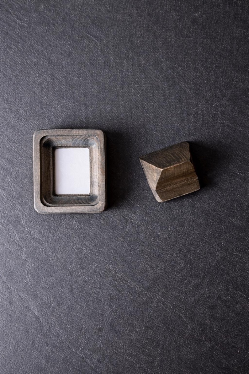 Puede incluir: Un peque&ntilde;o marco de fotos cuadrado de madera con un inserto blanco y un bloque de madera geom&eacute;trico. El marco tiene un acabado gris y un &aacute;rea empotrada para la foto. El bloque es de color marr&oacute;n oscuro y tiene un dise&ntilde;o facetado. Los objetos est&aacute;n sobre una superficie gris oscuro.