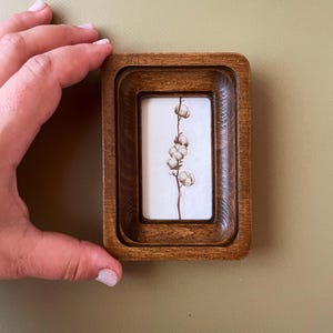 May include: A small, rectangular wooden frame with a watercolour painting of a cotton plant. The frame is a warm brown colour and has a layered design. The cotton plant has a thin, brown stem and several fluffy white cotton bolls.