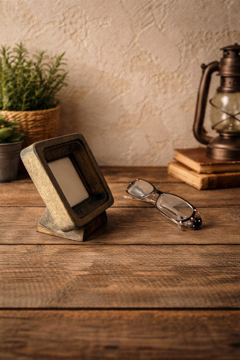 Puede incluir: Un marco de fotos de madera con un interior claro se encuentra sobre una superficie de madera. Gafas transparentes descansan cerca. La escena incluye una l&aacute;mpara de aceite vintage, libros y plantas en macetas, creando una est&eacute;tica r&uacute;stica.