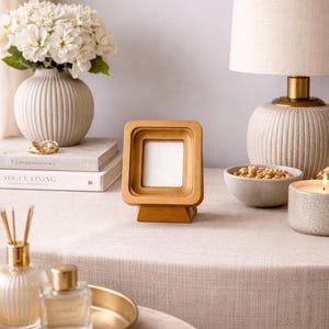 May include: A square wooden picture frame with a layered design, displayed on a table. The frame is light brown and has a white interior. The table is covered with a neutral-colored linen cloth, and other decorative items are visible.