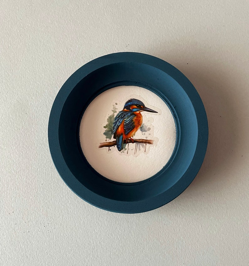 May include: A blue round frame with a white background featuring a watercolor painting of a kingfisher perched on a branch. The bird is blue, orange, and green with a long beak.