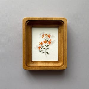 Könnte beinhalten: Ein kleiner, rechteckiger Holzrahmen mit einem Blumendruck. Der Druck zeigt orangefarbene Blumen mit grünen Stielen und Blättern. Der Rahmen ist aus hellem Holz gefertigt und hat ein schlichtes, elegantes Design. Das Kunstwerk ist zentriert.
