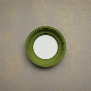 May include: A circular olive green frame with a smooth, matte finish and a wide, flat border. The frame features a recessed inner edge that creates depth, surrounding a plain, circular white center space. The design is simple and geometric.