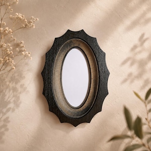 May include: An oval-shaped picture frame featuring a dark, textured black outer border with a scalloped edge and a smooth, muted gold-toned inner rim surrounding a plain white center panel, designed for wall mounting or display.