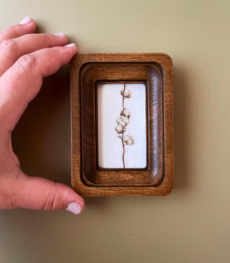 May include: A small, rectangular wooden frame with a watercolour painting of a cotton plant. The frame is a warm brown colour and has a layered design. The cotton plant has a thin, brown stem and several fluffy white cotton bolls.
