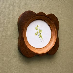 May include: A small, round, wooden frame with a scalloped edge. The frame holds a watercolor painting of delicate, yellow-green flowers on a white background. The frame is mounted on a muted green wall.