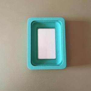May include: A rectangular teal wooden picture frame with rounded corners and a recessed center. The frame features a smooth, matte finish and a clean, simple design with a vertical orientation, surrounding a plain white rectangular insert.