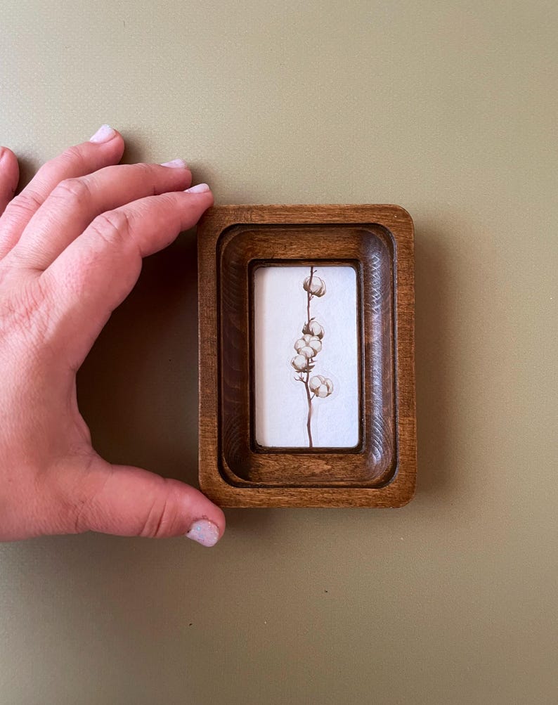 May include: A small, rectangular wooden frame with a recessed design. Inside, a white background displays a delicate botanical illustration of a cotton plant. The frame is held by a hand, showcasing the size and detail of the artwork.
