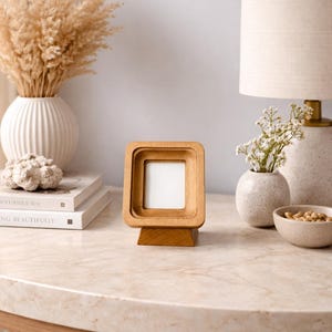 May include: A wooden picture frame with a square opening and a layered design. The frame is displayed on a light-colored table, with a lamp, vases, and books in the background. The frame is approximately 4 inches tall.