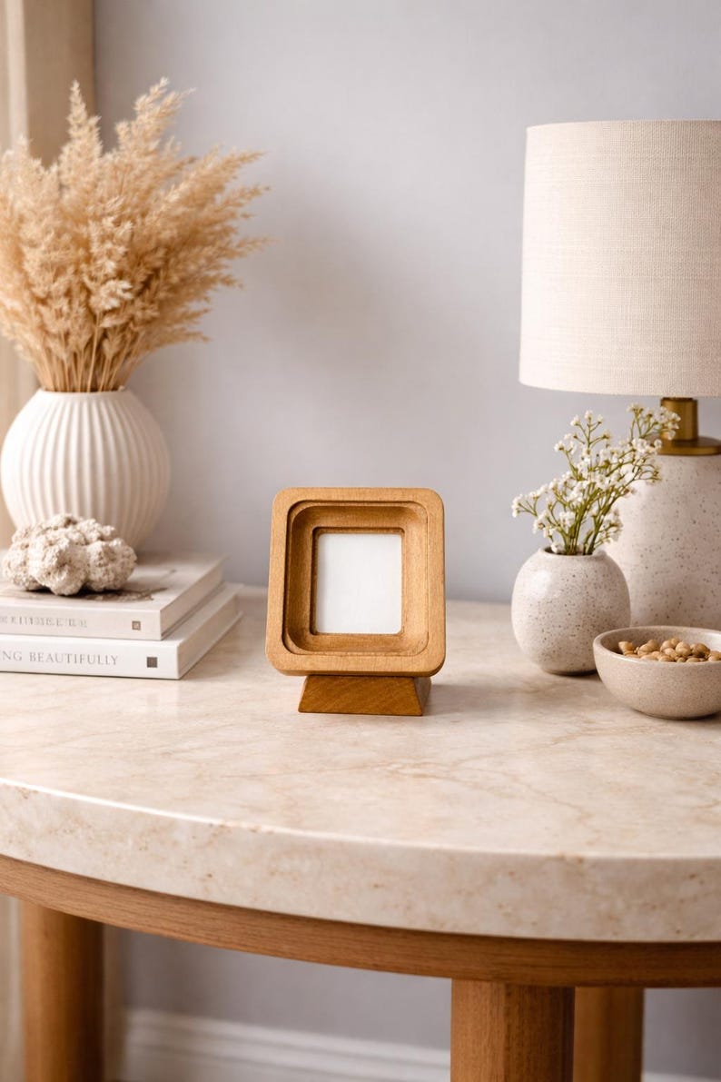 May include: A wooden picture frame with a square opening and a layered design. The frame is displayed on a light-colored table, with a lamp, vases, and books in the background. The frame is approximately 4 inches tall.