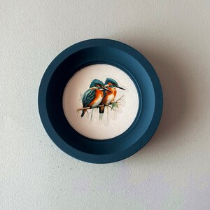 May include: A dark blue framed picture with a white background featuring a watercolor painting of two blue and orange birds perched on a branch.