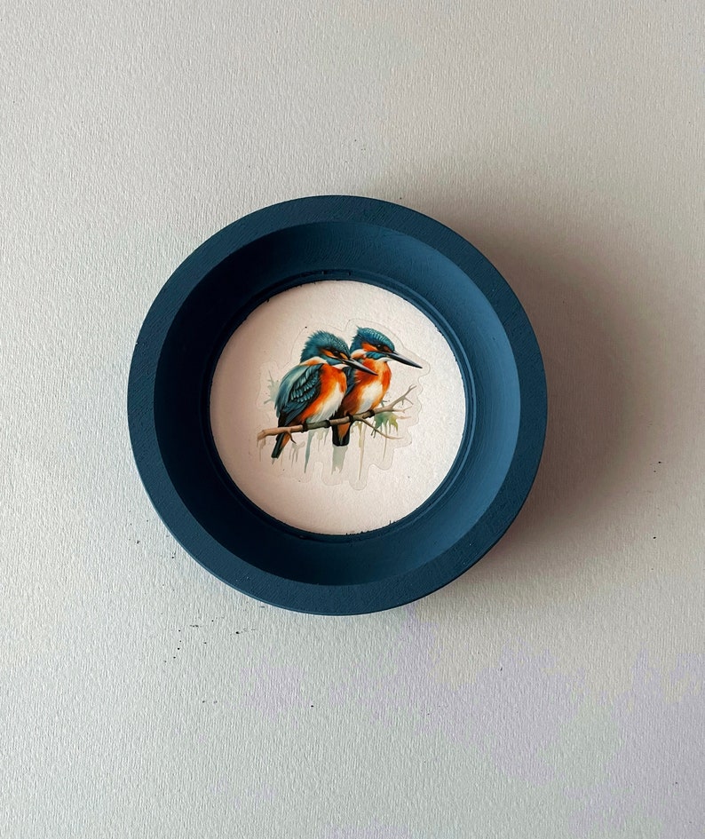 May include: A dark blue framed picture with a white background featuring a watercolor painting of two blue and orange birds perched on a branch.