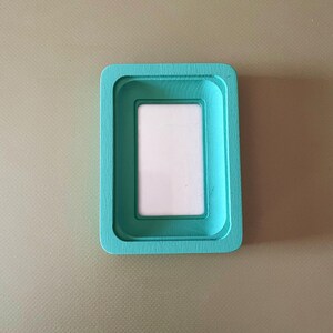 May include: A rectangular, teal-colored picture frame with a white interior. The frame has a layered design, with a recessed area for the photo. The frame is made of wood and has a smooth finish.