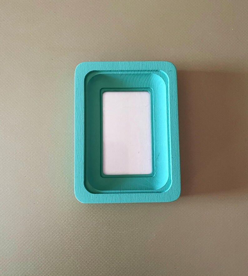 May include: A rectangular, teal-colored picture frame with a white interior. The frame has a layered design, with a recessed area for the photo. The frame is made of wood and has a smooth finish.