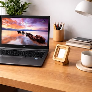 May include: A laptop with a colorful sunset wallpaper sits on a wooden desk, next to a small wooden picture frame. A white mug, pencils, and a desk lamp are also on the desk. A small green plant is in the background.