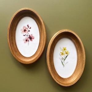 Pode incluir: Duas molduras ovais de madeira com arte botânica. Uma moldura apresenta flores cor-de-rosa, a outra flores amarelas. As molduras são montadas numa parede verde, criando uma exibição natural e artística.