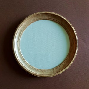 May include: A circular wooden picture frame with a light wood grain. The frame has a light blue interior and is set against a brown background. The frame is approximately 20 cm in diameter.