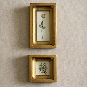 May include: Two gold-coloured framed botanical prints. The top frame is rectangular and contains a rose. The bottom frame is square and features a cluster of purple flowers. Both frames are mounted on a neutral-coloured wall.