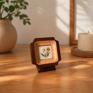 Pode incluir: Uma pequena moldura quadrada de madeira contendo um pedaço de tecido branco com um desenho floral bordado com flores brancas e amarelas e folhas verdes. A moldura tem um acabamento castanho escuro e está montada num suporte de madeira.