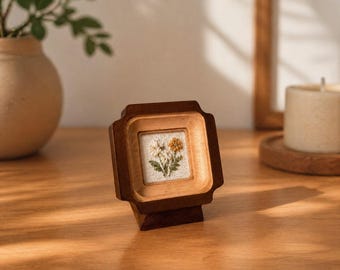May include: A small square wooden frame containing a piece of white fabric with an embroidered floral design featuring white and yellow flowers with green leaves. The frame has a dark brown finish and is mounted on a matching wooden stand.