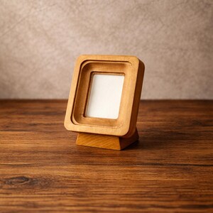 May include: A small, square wooden picture frame with a layered design. The frame is made of light-colored wood and has a white insert. It stands on a small wooden base, perfect for displaying a photograph or artwork.