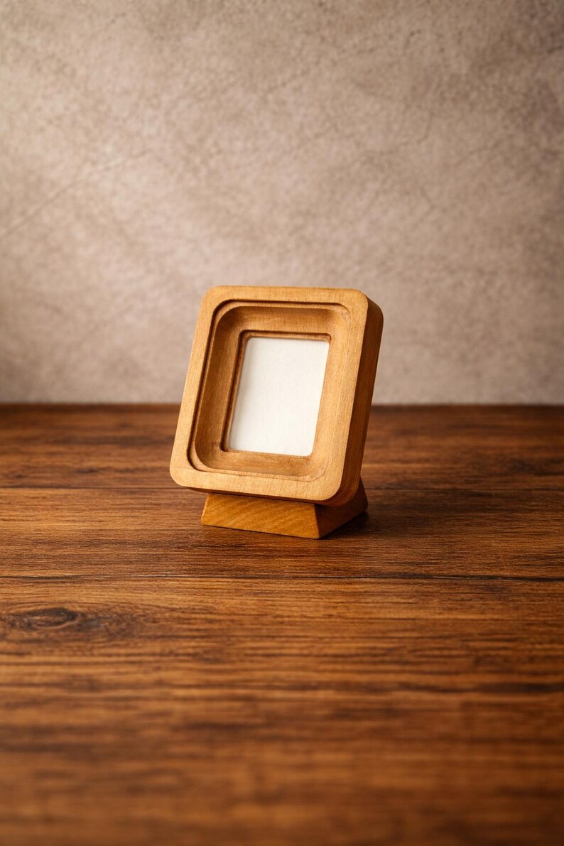 May include: A small, square wooden picture frame with a layered design. The frame is made of light-colored wood and has a white insert. It stands on a small wooden base, perfect for displaying a photograph or artwork.