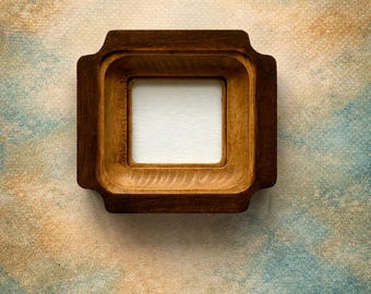 May include: A square wooden frame with a dark brown finish and a smooth, recessed inner border. The frame features a distinct stepped design with rounded corners on the outer edges, surrounding a plain, white, square-shaped center panel.