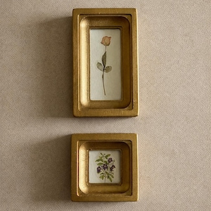 Pode incluir: Duas pequenas obras de arte botânica emolduradas em dourado. A moldura superior contém uma rosa prensada, e a moldura inferior apresenta um grupo de flores roxas. As molduras são retangulares com um design ligeiramente curvo e rebaixado.