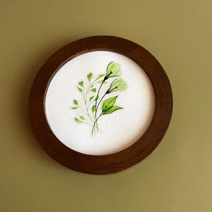 May include: A round, dark brown wooden frame surrounds a watercolor painting of green leaves and flowers on a white background. The artwork features delicate, translucent green floral elements and vibrant green leaves, creating a natural, botanical theme.