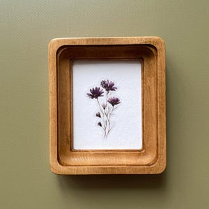 May include: A small, rectangular wooden frame with a light brown finish. Inside the frame is a white background with a cluster of purple flowers. The frame is set against a muted green background.
