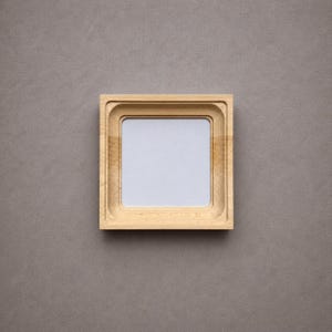 Pode incluir: Uma moldura quadrada de madeira com um acabamento de grão natural em tom bronzeado claro. A moldura apresenta uma borda interna escalonada e rebaixada que leva a um painel central quadrado de cor clara, oferecendo uma superfície simples.