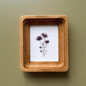 May include: A small, rectangular wooden frame with a light brown finish. Inside the frame is a white background with a cluster of dark purple flowers. The frame is set against a muted green background.