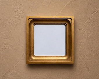 May include: A square picture frame with a metallic gold finish and a stepped, recessed inner border. The frame features a smooth, reflective surface and contains a plain, white, square-shaped centre insert for displaying artwork or photographs.
