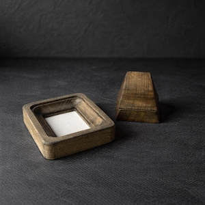 Puede incluir: Un marco de fotos de madera con un inserto blanco y un peque&ntilde;o objeto geom&eacute;trico de madera. El marco es cuadrado con un &aacute;rea empotrada para la foto. Los objetos son de madera de color marr&oacute;n claro y est&aacute;n sobre una superficie oscura.